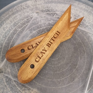 May include: Two wooden pottery tools, each engraved with "CLAY BITCH". The tools are light brown, with a pointed end and a hole. They are resting on a clear plastic surface, likely a potter's wheel.