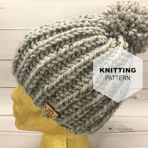 Könnte beinhalten: Eine graue Strickmütze mit einem großen Bommel oben. Die Mütze ist aus einem dicken Garn gestrickt und hat ein einfaches Stichmuster. Die Mütze befindet sich auf einem Mannequin-Kopf und der Text "KNITTING PATTERN" ist auf dem Bild zu sehen.