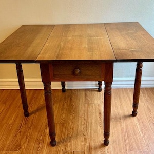 Puede incluir: Una mesa de madera con hojas abatibles y un acabado marrón medio cálido. La mesa tiene un solo cajón con un pomo redondo de madera y cuatro patas torneadas. Las hojas están extendidas, creando una superficie más grande. La mesa mide unos 76 cm de alto.