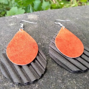 May include: A pair of handmade earrings featuring a layered design. The top layer is a teardrop shape in a vibrant orange, while the bottom layer is a textured, dark gray, semi-circular shape. The earrings have silver-colored hooks.