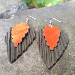 May include: A pair of dangle earrings with a layered design. The top layer is a bright orange suede leaf shape, and the bottom layer is a dark gray textured leaf shape. The earrings are attached to silver hooks.