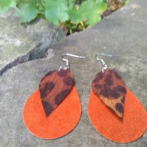 May include: A pair of dangle earrings with orange suede and brown leopard print fabric. The earrings are hanging from silver hooks.