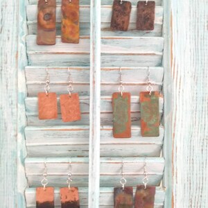 May include: Six pairs of rectangular copper earrings with different patterns and colors displayed on a white wooden shutter.