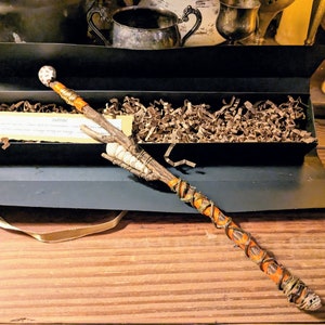 May include: A wooden wand with a white and brown orb on the end. The wand is wrapped in brown leather and has a natural branch design. The wand is in a black box with brown paper shredding.