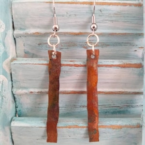 May include: A pair of silver dangle earrings with long, thin, rusty brown metal pieces hanging from them. The earrings are hanging on a light blue wooden surface.