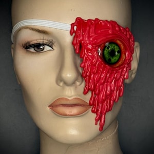 May include: A mannequin head with a red, textured eye patch covering one eye. The eye patch has a dripping wax-like appearance and features a green and brown eye. A white elastic band secures the patch.