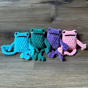 May include: Four crocheted frog plushies in different colors: teal, green, purple, and pink. The frogs are sitting on a wooden surface.