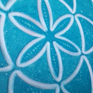 May include: A close-up of a teal blue background with a white, stylized, geometric design. The design resembles a flower with multiple petals.