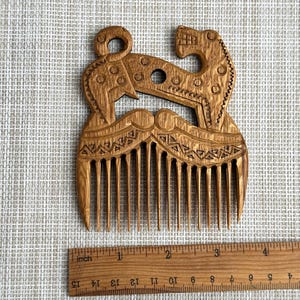 May include: A light brown wooden hair comb featuring an intricate carved design of two stylized animals facing each other. The comb is positioned above a wooden ruler, showing the object is approximately 3.5 inches wide at its widest point.
