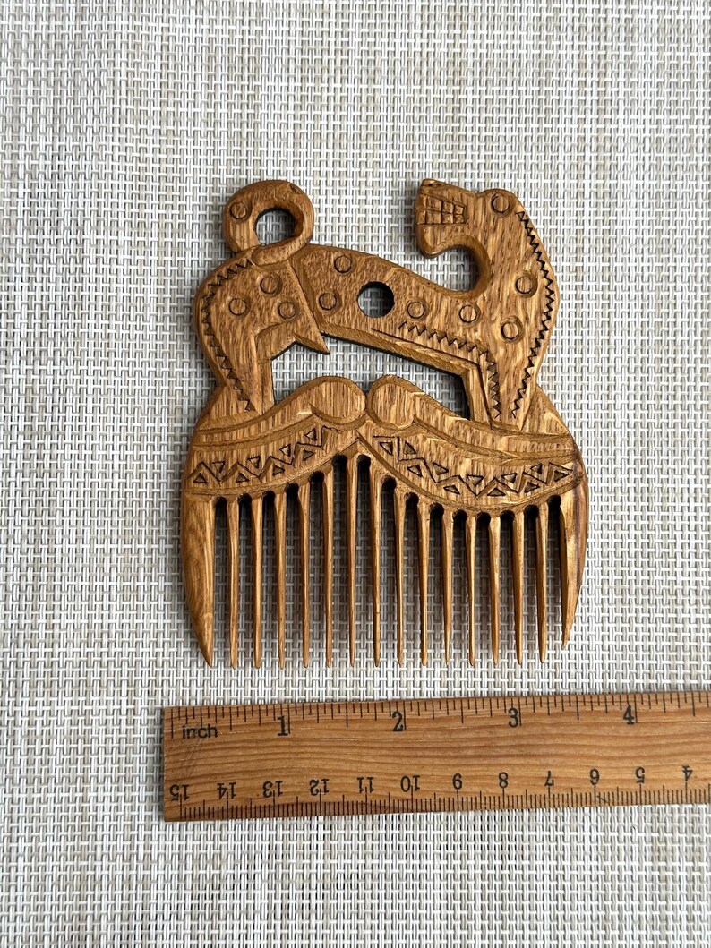 May include: A light brown wooden hair comb featuring an intricate carved design of two stylized animals facing each other. The comb is positioned above a wooden ruler, showing the object is approximately 3.5 inches wide at its widest point.