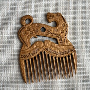 May include: A light brown wooden comb featuring an intricate carved design of a stylized animal with a curled tail and open mouth. The handle area displays geometric patterns, including triangles and circles, above eleven vertical teeth.