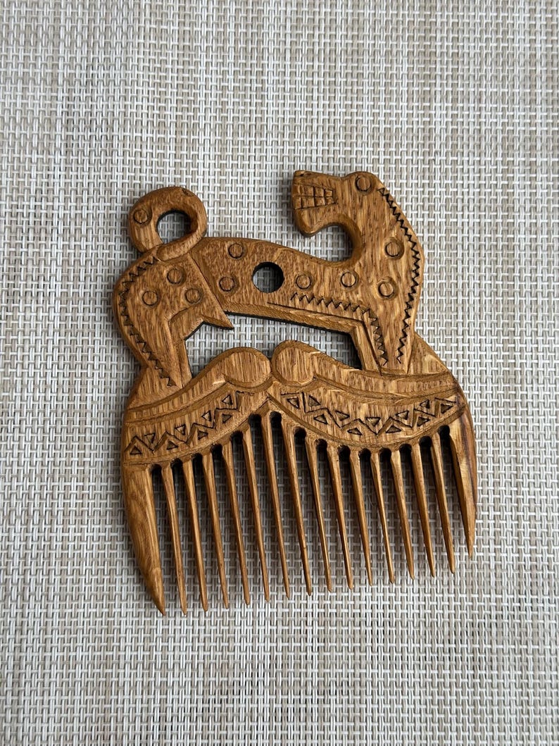 May include: A light brown wooden comb featuring an intricate carved design of a stylized animal with a curled tail and open mouth. The handle area displays geometric patterns, including triangles and circles, above eleven vertical teeth.