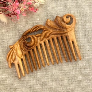 Pode incluir: Um pente de cabelo de madeira castanho-claro com um intrincado design floral esculpido ao longo da borda superior. O pente tem onze dentes lisos e cónicos com um acabamento polido que realça os padrões naturais dos veios da madeira.