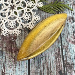 Puede incluir: Un cuenco de madera para lana con un acabado de veta de madera natural. El cuenco es de color amarillo claro con un patrón de veta de madera marrón oscuro. El cuenco tiene forma de óvalo largo con una ligera curva en la parte superior.