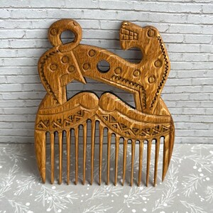 May include: A light brown wooden hair comb featuring an intricate carved design of a stylized animal with a long neck and tail. The handle area displays geometric patterns, including circles and triangles, etched into the wood grain surface.