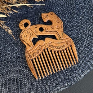 May include: A light brown wooden hair comb featuring an intricate carved design of a stylized animal with geometric patterns. The comb has a series of fine teeth at the bottom and a decorative handle shaped like a creature with circular details.