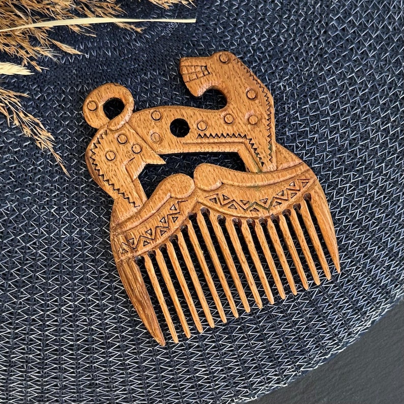 May include: A light brown wooden hair comb featuring an intricate carved design of a stylized animal with geometric patterns. The comb has a series of fine teeth at the bottom and a decorative handle shaped like a creature with circular details.