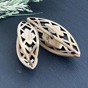 May include: Two wooden, oval-shaped earrings with intricate cut-out designs. The earrings are a light brown color and have a geometric pattern. The earrings are displayed on a dark surface.