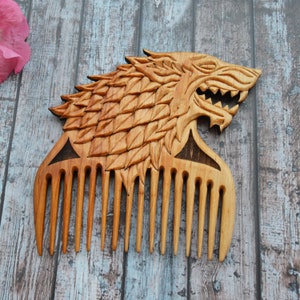 May include: A wooden comb carved in the shape of a wolf's head. The comb has a brown wood finish and features a detailed carving of the wolf's face and fur. The comb has 10 teeth.