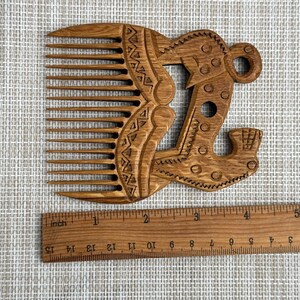 May include: A brown wooden comb with long teeth and an ornate handle featuring carved geometric patterns and circular motifs. A wooden ruler is positioned below the comb, showing the object is approximately 5.5 inches in width.