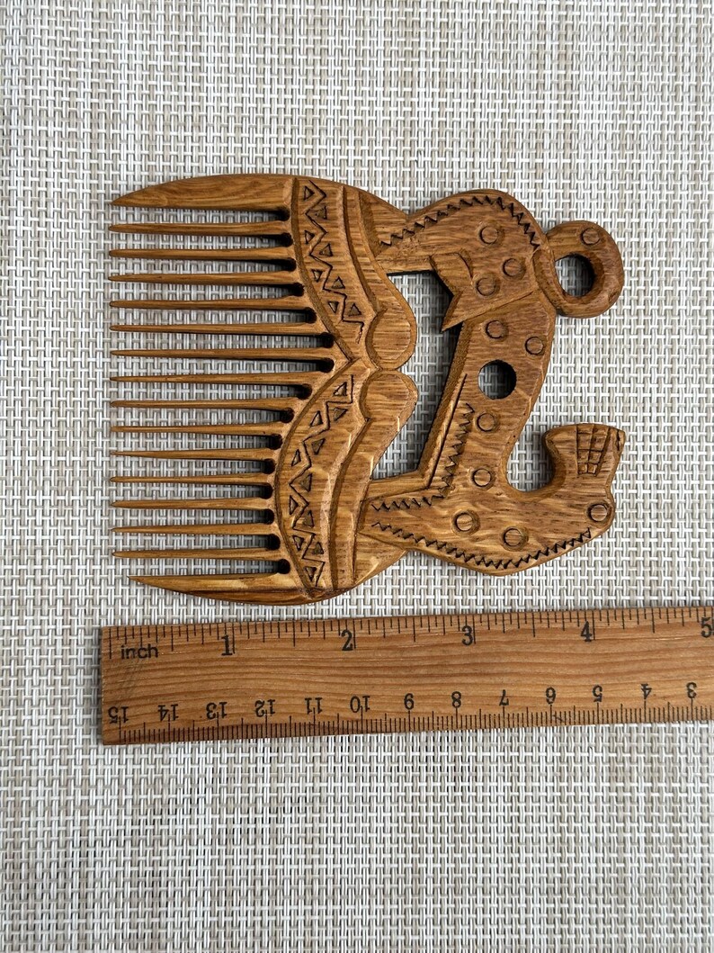 May include: A brown wooden comb with long teeth and an ornate handle featuring carved geometric patterns and circular motifs. A wooden ruler is positioned below the comb, showing the object is approximately 5.5 inches in width.