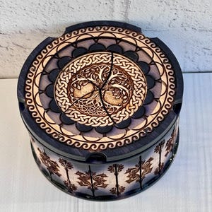 Puede incluir: Una caja redonda de madera con un diseño celta del árbol de la vida en la tapa. La caja es azul oscuro con intrincados detalles tallados en un color de madera más claro. Los lados presentan un patrón floral repetido.