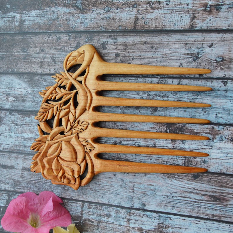 May include: A wooden comb with a carved rose design. The comb is made of light brown wood and has seven teeth. The rose is carved in detail and has a realistic appearance.