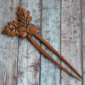 May include: A wooden hair fork with an acorn and oak leaf design. The fork is brown and has two prongs.