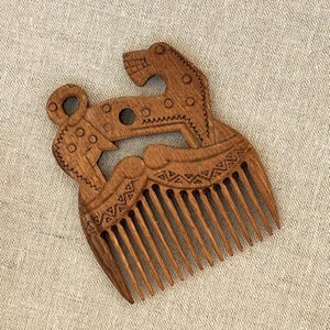 May include: A brown wooden hair comb featuring an intricate carved design of a stylized animal figure. The handle area displays geometric patterns, circular cutouts, and linear engravings, while the lower section consists of eleven long, thin teeth.