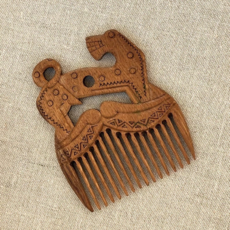 May include: A brown wooden hair comb featuring an intricate carved design of a stylized animal figure. The handle area displays geometric patterns, circular cutouts, and linear engravings, while the lower section consists of eleven long, thin teeth.