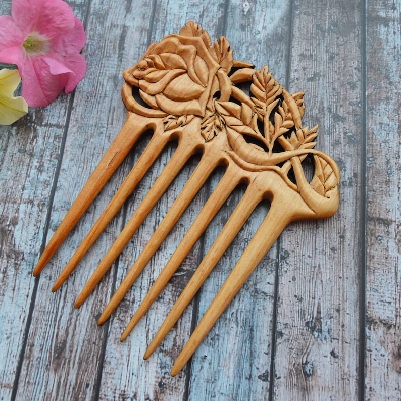 May include: A wooden hair comb with a detailed carving of roses and leaves. The comb has seven teeth and is made of light brown wood.