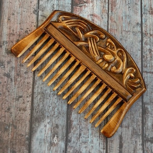 May include: A wooden comb with a carved design of a knotwork pattern. The comb is brown and has a natural finish.