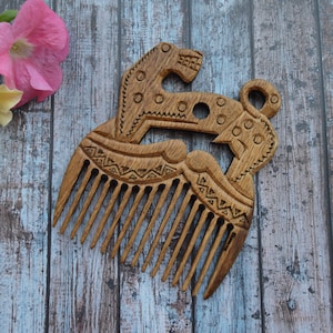 May include: A wooden comb with an intricate carved design featuring two animal figures. The comb has a series of teeth and is decorated with geometric patterns and circular accents. The comb is a light brown color.