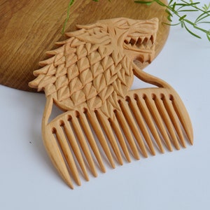 May include: A wooden comb carved in the shape of a wolf's head. The comb has a detailed design and is made of light brown wood.