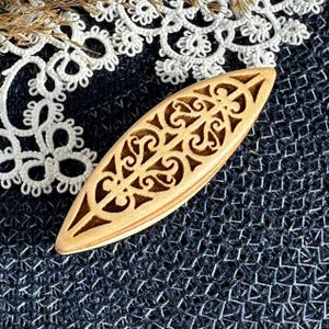 May include: A light brown wooden hair accessory with a pointed, elongated oval shape and intricate, symmetrical cut-out scrollwork patterns throughout the surface, showcasing a natural wood grain texture and a smooth, polished finish.