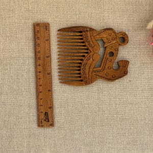 May include: A brown wooden hair comb with carved geometric patterns and a stylized animal figure, positioned next to a wooden ruler showing a length of approximately 6 inches for the comb's width and height dimensions.
