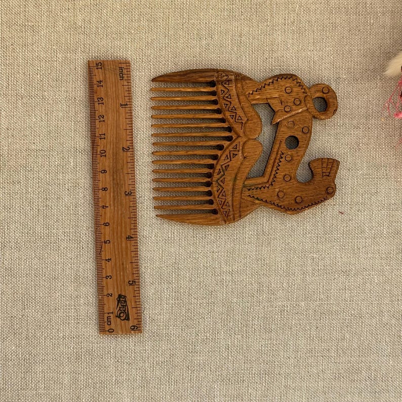 May include: A brown wooden hair comb with carved geometric patterns and a stylized animal figure, positioned next to a wooden ruler showing a length of approximately 6 inches for the comb's width and height dimensions.