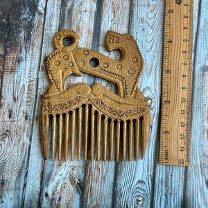 May include: A light brown wooden hair comb featuring an intricate carved design of two stylized animals facing each other, positioned next to a wooden ruler that shows the comb is approximately 3.5 inches wide and 4 inches in total height.