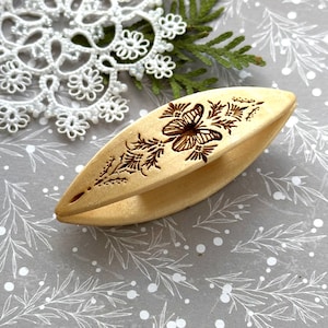 May include: A wooden crochet hook with a butterfly and floral design carved into the surface. The hook is light brown and has a smooth finish.