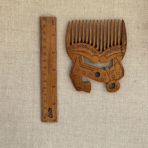May include: A brown wooden hair comb with a wide-tooth design and intricate etched patterns, positioned next to a wooden ruler that shows the comb is approximately 6 inches in height and 4.5 inches in width at its widest point.