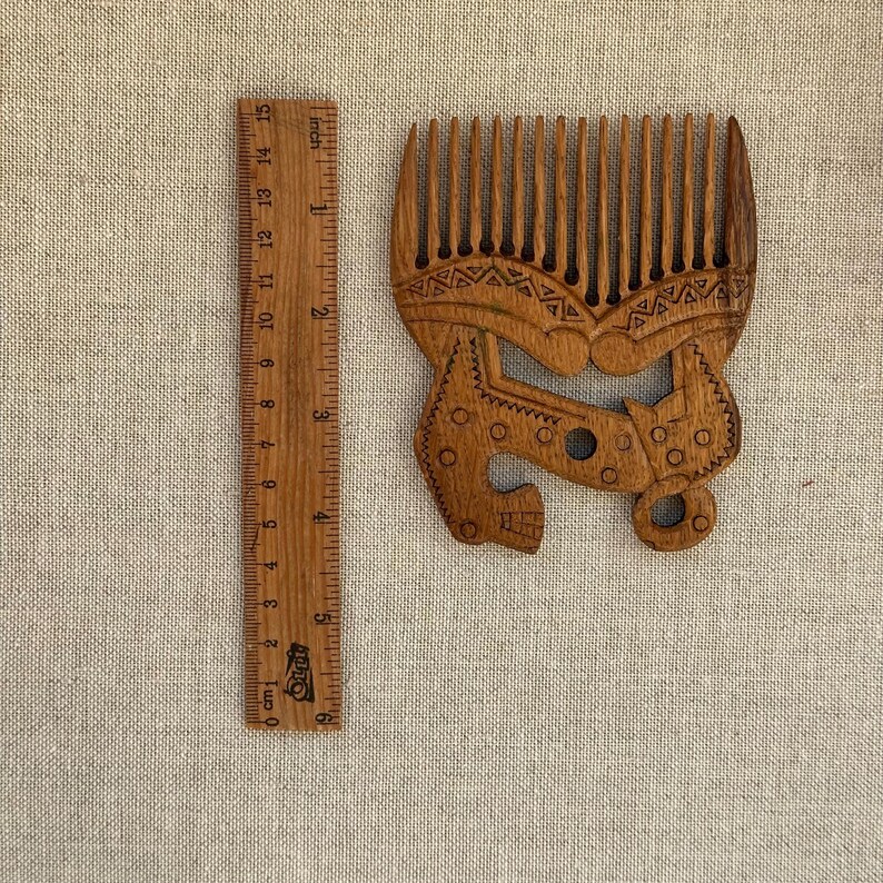 May include: A brown wooden hair comb with a wide-tooth design and intricate etched patterns, positioned next to a wooden ruler that shows the comb is approximately 6 inches in height and 4.5 inches in width at its widest point.