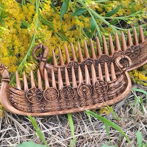 May include: Two intricately carved wooden combs, shaped like Viking longboats, rest on a bed of dry grass and yellow flowers. The combs are brown with detailed carvings of shields and Viking ship features. The combs are approximately 6 inches long.