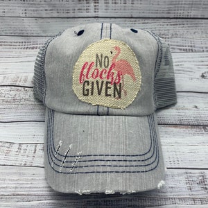 May include: A gray and blue mesh trucker hat with a distressed look. The hat has a patch with a pink flamingo and the text "No flocks given".