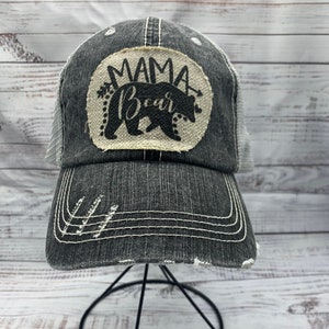 May include: A black and white distressed trucker hat with a white mesh back. The hat has a patch on the front that says "Mama Bear" with a black bear silhouette.