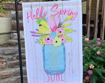 May include: A white garden flag with a blue mason jar filled with colorful flowers and the text "Hello Spring" in pink lettering.