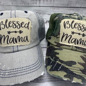 May include: Two trucker hats with a distressed look. One hat is gray with a blue mesh back and the other is camouflage with a green mesh back. Both hats have a square patch with the text "Blessed Mama" and an arrow.