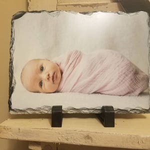 May include: A photo of a baby wrapped in a pink blanket, printed on a slate stone with a stand.