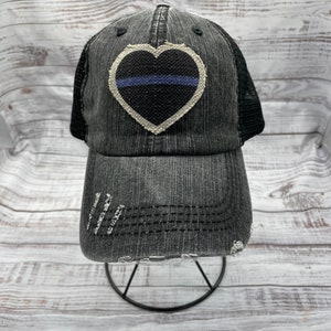 May include: A black and white distressed denim trucker hat with a black heart-shaped patch featuring a thin blue line. The hat has a mesh back.