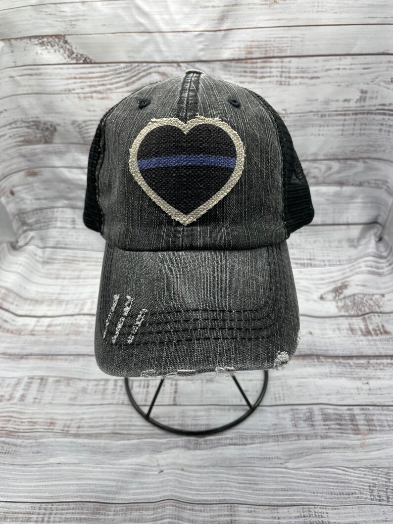 May include: A black and white distressed denim trucker hat with a black heart-shaped patch featuring a thin blue line. The hat has a mesh back.