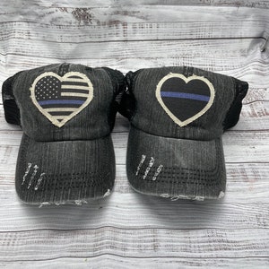 May include: Two black denim trucker hats with distressed detailing. One hat features a white heart with an American flag and a blue line, the other hat features a white heart with a blue line.